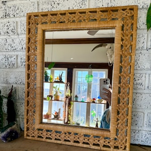 May include: Rectangular woven rattan mirror with a decorative braided border. The mirror reflects a room with windows, plants, and a shelf with various decorative items. The frame is a light tan color.