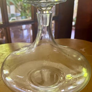 May include: A clear glass decanter featuring a wide, rounded base and a slender neck that tapers upward. The vessel is topped with a flat, circular glass stopper that sits flush with the opening, creating a smooth, continuous silhouette.