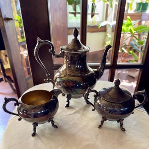 May include: A silver tea set with a teapot, creamer, and sugar bowl. The teapot has a curved handle and a decorative finial on the lid. The creamer and sugar bowl have matching ornate handles and sit on three small legs. The set is displayed on a white tablecloth.