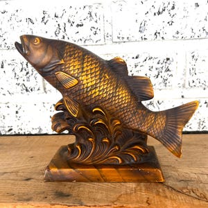 May include: A wooden sculpture of a fish jumping out of the water. The fish is painted in shades of brown and gold. The sculpture is on a wooden base.