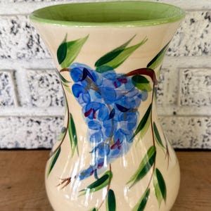May include: A ceramic vase with a cream-colored base, a green rim, and a green interior. The vase is decorated with a blue floral design and green leaves. The vase is sitting on a wooden surface.