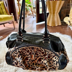 May include: A black handbag with a textured crocodile-skin pattern and a front pocket featuring a leopard print design. The bag includes two long shoulder straps, silver-toned hardware, and a small metallic padlock attached to the front zipper.