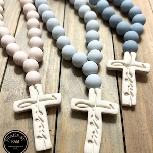 May include: Three rosaries with large, round beads in shades of pink, light blue, and blue. Each rosary has a white cross pendant with etched text. The rosaries are displayed on a wooden surface.