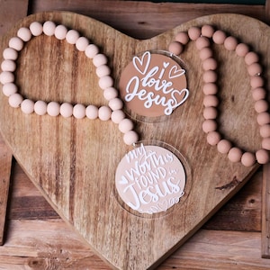 May include: A wooden heart-shaped tray holds two round, clear acrylic ornaments with inspirational messages. One says "I love Jesus" and the other says "My Worth is Found in Jesus." A beaded garland in a light pink hue surrounds the ornaments.