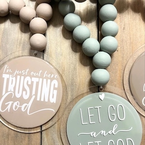 May include: Two decorative hanging ornaments featuring strings of wooden beads in beige and sage green. Each ornament has a clear circular acrylic disc with white text. One disc reads I'm just out here TRUSTING God, and the other reads LET GO and LET GOD with a small white heart icon.