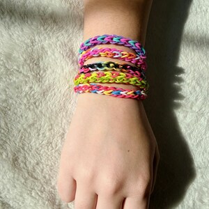 May include: A stack of colorful rubber band bracelets in various shades of pink, blue, yellow, green, and black. The bracelets are worn on a wrist, showcasing the woven design and vibrant colors.