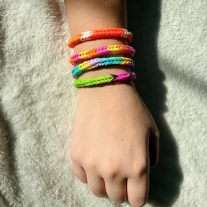 May include: A collection of colorful loom bracelets stacked on a wrist. The bracelets feature a variety of bright colors, including orange, pink, yellow, green, and blue. Each bracelet is made of woven rubber bands.