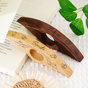 May include: Two wooden thumb page holders, one light and one dark, with floral and butterfly engravings. The light-colored holder has the name "Bonita" engraved. The dark-colored holder has the name "Nafisa" engraved.