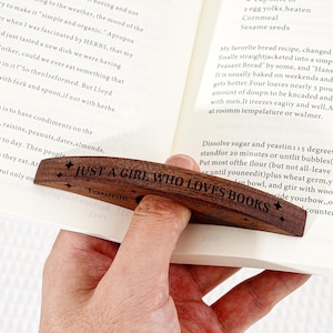 May include: A wooden book page holder with the text "JUST A GIRL WHO LOVES BOOKS" engraved on it. The dark brown holder has a curved design and is held open on a book. The holder is approximately 4 inches long.
