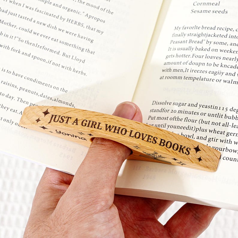 May include: Wooden book page holder with the text "JUST A GIRL WHO LOVES BOOKS" and star designs. The light brown holder is held in place by a finger, keeping the book open. The book is open to a page of text.