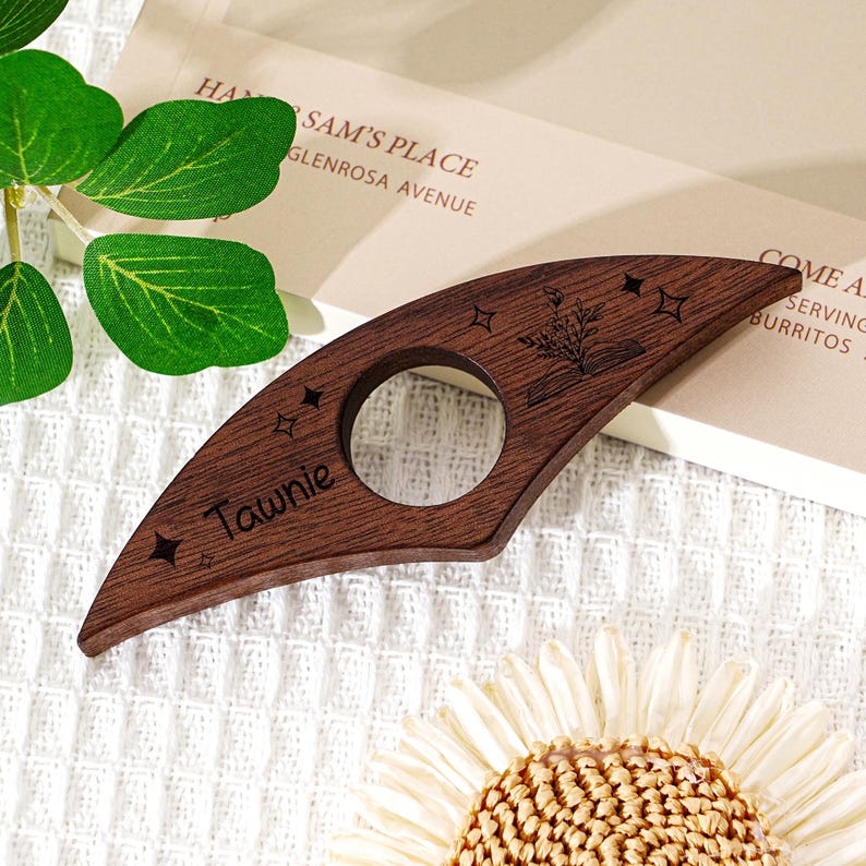 May include: A dark brown wooden page holder with a circular opening and a pointed end. The page holder is engraved with the name "Tawnie", stars, and a floral design. The background includes a book and a flower.