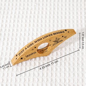 May include: A wooden book page holder with the text "JUST A GIRL WHO LOVES BOOKS" and the name "Yonina". The holder is 4.33 inches (11 cm) long and 1.37 inches (3.5 cm) wide. It features a cutout for the thumb and decorative stars.