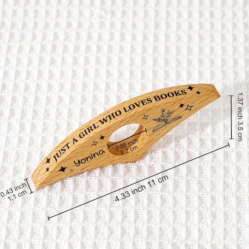 May include: A wooden book page holder with the text "JUST A GIRL WHO LOVES BOOKS" and the name "Yonina". The holder is 4.33 inches (11 cm) long and 1.37 inches (3.5 cm) wide. It features a cutout for the thumb and decorative stars.