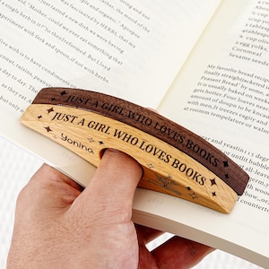 May include: A wooden book page holder with the text "JUST A GIRL WHO LOVES BOOKS" engraved on it. The holder is light brown with a dark brown accent and has a finger hole for easy use. The name "Yonina" is also engraved.