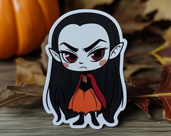 May include: A sticker featuring a cartoon vampire with long black hair, red eyes, and a stern expression. The vampire wears an orange dress and a red cape. The background includes a pumpkin and autumn leaves.