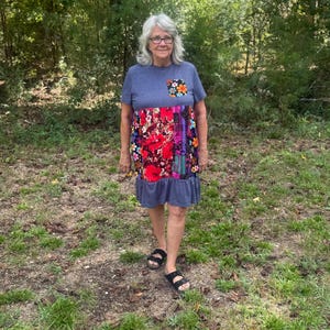 Puede incluir: Una mujer con un vestido azul con un diseño de patchwork floral en el corpiño y un dobladillo con volantes. El vestido tiene un pequeño bolsillo con un diseño floral. Lleva sandalias negras y está de pie en una zona cubierta de hierba.