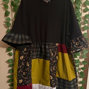 Puede incluir: Vestido patchwork negro con cuello redondo y mangas cortas. El vestido presenta una mezcla de telas estampadas, incluyendo flores, cuadros y colores lisos como amarillo mostaza y burdeos. Las mangas tienen puños con volantes.