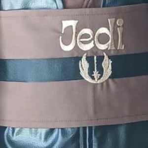 May include: A wide brown fabric belt featuring a horizontal black stripe across the center. The word Jedi is embroidered in light-colored thread above a circular emblem with wings and a central star, both stitched in a matching light-colored thread.