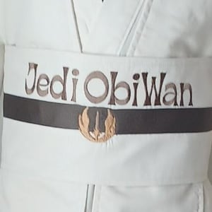 May include: A white fabric sash featuring the embroidered text Jedi ObiWan in brown lettering. A horizontal dark brown band runs across the center, accented with a gold-colored embroidered Jedi Order symbol centered below the text.