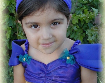 May include: A young girl wearing a purple costume with a blue jeweled headband. The costume has a gathered bodice with gold fringe and a matching sash. The girl is smiling.