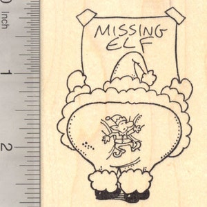 May include: A rubber stamp featuring a black line drawing of a sign that reads MISSING ELF, with a smaller elf illustration below. A ruler on the left shows the stamp is approximately 5 cm tall. The design is on a light wood block.