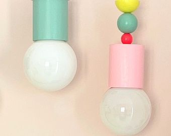 Op de afbeelding: Twee hangende lampen met kleurrijke geometrische vormen en witte gloeilampen. De lamp aan de linkerkant heeft een lichtgroene cilinder, een roze bol, een blauwe kom en een gele bol. De lamp aan de rechterkant heeft een roze cilinder, een rode bol, een groene bol en een gele kom.