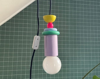 Op de afbeelding: Een kleurrijke hanglamp met een geometrisch ontwerp in paars, groen, blauw, geel en roze. De lamp hangt aan een zwart snoer en heeft een witte gloeilamp.