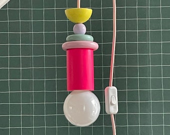 Op de afbeelding: Een roze, gele, groene en paarse hanglamp met een witte gloeilamp die hangt aan een roze snoer tegen een groene rasterachtergrond.