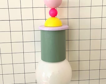 Op de afbeelding: Een groene en witte hanglamp met een geometrisch ontwerp in roze, geel en rood. De lamp hangt aan een wit plafond.