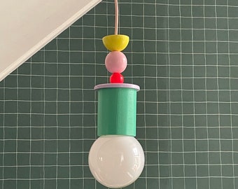 Op de afbeelding: Een groene hanglamp met een witte gloeilamp. De lamp heeft een stapel kleurrijke geometrische vormen: een gele halve cirkel, een roze bol, een rode bol en een paarse halve cirkel.