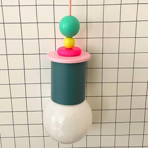Peut inclure: Un luminaire suspendu moderne et coloré. Il se compose d'une ampoule blanche, d'un cylindre turquoise, d'un disque rose et de sphères empilées vertes, jaunes et roses. Le luminaire est suspendu à un cordon rose.