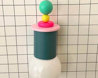 Op de afbeelding: Een moderne, kleurrijke hanglamp. De lamp heeft een witte lampvoet, een teal cilinder, een roze schijf en gestapelde groene, gele en roze bollen. De lamp hangt aan een roze snoer.