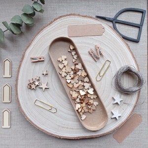 May include: A wooden tray filled with a selection of wooden heart shapes, stars, and other shapes. The tray is on a wooden surface with a brown paper tag, a pair of scissors, and a spool of twine.