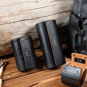 May include: Black leather watch cases with a crocodile texture are displayed on a wooden surface. The cases are cylindrical and rectangular, with personalized initials in gold. A black backpack is in the background, and a watch case is open.