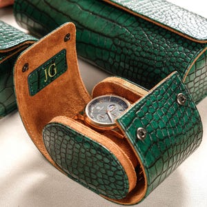 Puede incluir: Un estuche cilíndrico para reloj con exterior de piel sintética verde oscuro con textura de cocodrilo e interior de ante color canela. El estuche está abierto, mostrando un reloj de pulsera redondo con marco metálico y esfera oscura. La solapa muestra JG.