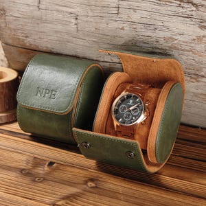 May include: A green leather watch case with the initials "NPE" embossed on the lid. The case is open, revealing a gold-toned watch with a black face. The watch has multiple subdials and a gold band. The interior of the case is a tan color.