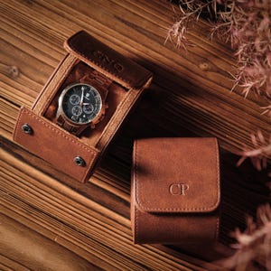 K&ouml;nnte beinhalten: Ein braunes Uhrenetui aus Leder mit Druckknopfverschluss, das eine Chronographenuhr mit dunklem Zifferblatt und Metallarmband enth&auml;lt. Das Etui ist mit den Buchstaben CNQ gepr&auml;gt. Ein zweites geschlossenes braunes Lederetui tr&auml;gt die Buchstaben CP.