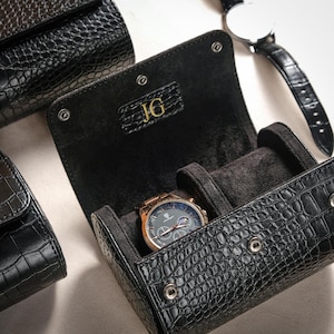 May include: Black leather watch roll with a crocodile texture, holding a gold and gray watch. The roll is open, revealing a soft interior and the initials "JG" in gold. Other closed watch rolls are visible in the background.