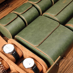 K&ouml;nnte beinhalten: Eine Sammlung von Uhrenaufbewahrungsboxen aus olivgr&uuml;nem Leder mit Klappverschl&uuml;ssen und beigefarbenem Innenfutter. Mehrere Etuis tragen gepr&auml;gte Initialen wie SP und NPW. Zwei Armbanduhren mit wei&szlig;em Zifferblatt sind zu sehen.