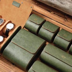 K&ouml;nnte beinhalten: Eine Sammlung von olivgr&uuml;nen Uhrenaufbewahrungsetuis aus Leder mit Klappverschl&uuml;ssen und goldgepr&auml;gten Initialen wie SP, NPE, SG und WP. Das Set enth&auml;lt ein gr&ouml;&szlig;eres offenes Etui, in dem zwei Armbanduhren mit wei&szlig;em Zifferblatt liegen.