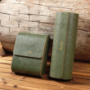 May include: Two green leather cases, one rectangular with the initials "AIW" and the other cylindrical with the word "Brave" in gold lettering. The cases are on a wooden surface with a wooden background.