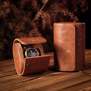 K&ouml;nnte beinhalten: Zwei zylindrische braune Uhrenetuis mit Druckknopfverschl&uuml;ssen. Ein Etui ist ge&ouml;ffnet und zeigt eine Armbanduhr mit schwarzem Zifferblatt und silbernen Zeigern. Das offene Etui tr&auml;gt die Pr&auml;gung CNQ, das geschlossene Etui zeigt goldene Buchstaben BDP.