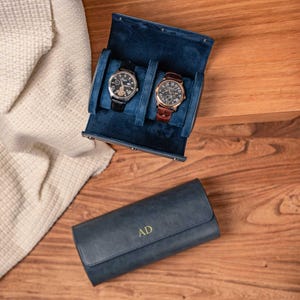May include: A navy blue leather watch roll case with the gold letters AD embossed on the front. The open case features a dark blue velvet interior holding two wristwatches, one with a black strap and one with a brown leather strap.