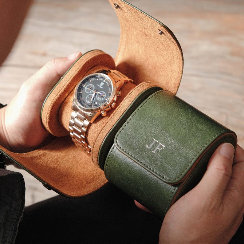 K&ouml;nnte beinhalten: Ein zylindrisches Uhrenetui in Waldgr&uuml;n mit beigefarbenem Innenfutter und den eingepr&auml;gten Initialen JF auf der Au&szlig;enklappe. Das Etui ist ge&ouml;ffnet und zeigt eine silberfarbene Chronographen-Armbanduhr mit schwarzem Zifferblatt und Metallarmband.