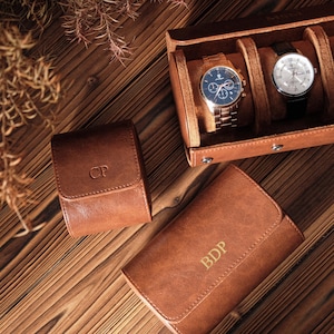 K&ouml;nnte beinhalten: Ein Set aus braunen Lederetuis zur Uhrenaufbewahrung mit personalisierten Monogrammen. Ein Etui ist ge&ouml;ffnet und zeigt zwei Armbanduhren mit Metallarmb&auml;ndern und runden Zifferbl&auml;ttern. Zwei weitere geschlossene Etuis tragen die Buchstaben CP und BDP.