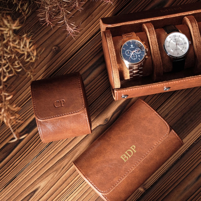 K&ouml;nnte beinhalten: Ein Set aus braunen Lederetuis zur Uhrenaufbewahrung mit personalisierten Monogrammen. Ein Etui ist ge&ouml;ffnet und zeigt zwei Armbanduhren mit Metallarmb&auml;ndern und runden Zifferbl&auml;ttern. Zwei weitere geschlossene Etuis tragen die Buchstaben CP und BDP.