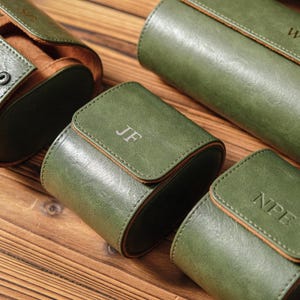 K&ouml;nnte beinhalten: Eine Sammlung von Uhrenrollen aus olivgr&uuml;nem Leder mit hellbraunem Innenfutter und Druckkn&ouml;pfen. Jede Rolle weist ein einzigartiges gepr&auml;gtes Monogramm auf, darunter die Buchstaben JF und NPE, mit sichtbaren N&auml;hten an den Kanten der abgerundeten Klappen.
