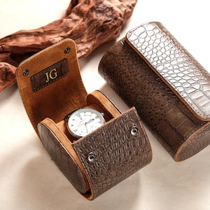 May include: Brown leather watch case with a crocodile pattern. The case is open, revealing a silver watch with a white face. The initials "JG" are embossed on the inside flap. Another closed case is visible.