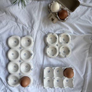 May include: White ceramic egg holders with blue dot detailing. The image displays three different styles of egg holders, each holding one or more brown eggs. A cardboard egg box with two eggs is also visible. The egg holders are on a white fabric surface.