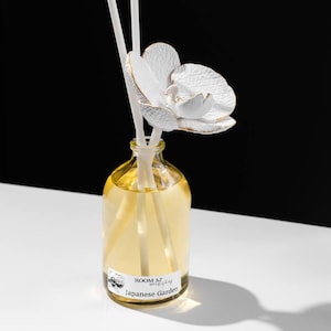 May include: A clear glass bottle diffuser with a white ceramic orchid on top. The bottle is filled with a yellow liquid and has a label that reads "ROOM 3.7  workshop  Japanese Garden".